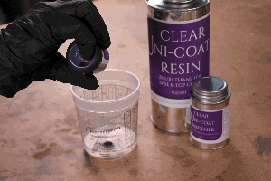 gloved hand pouring violet tint into clear measuring cup with Uni-coat urethane resin next to it